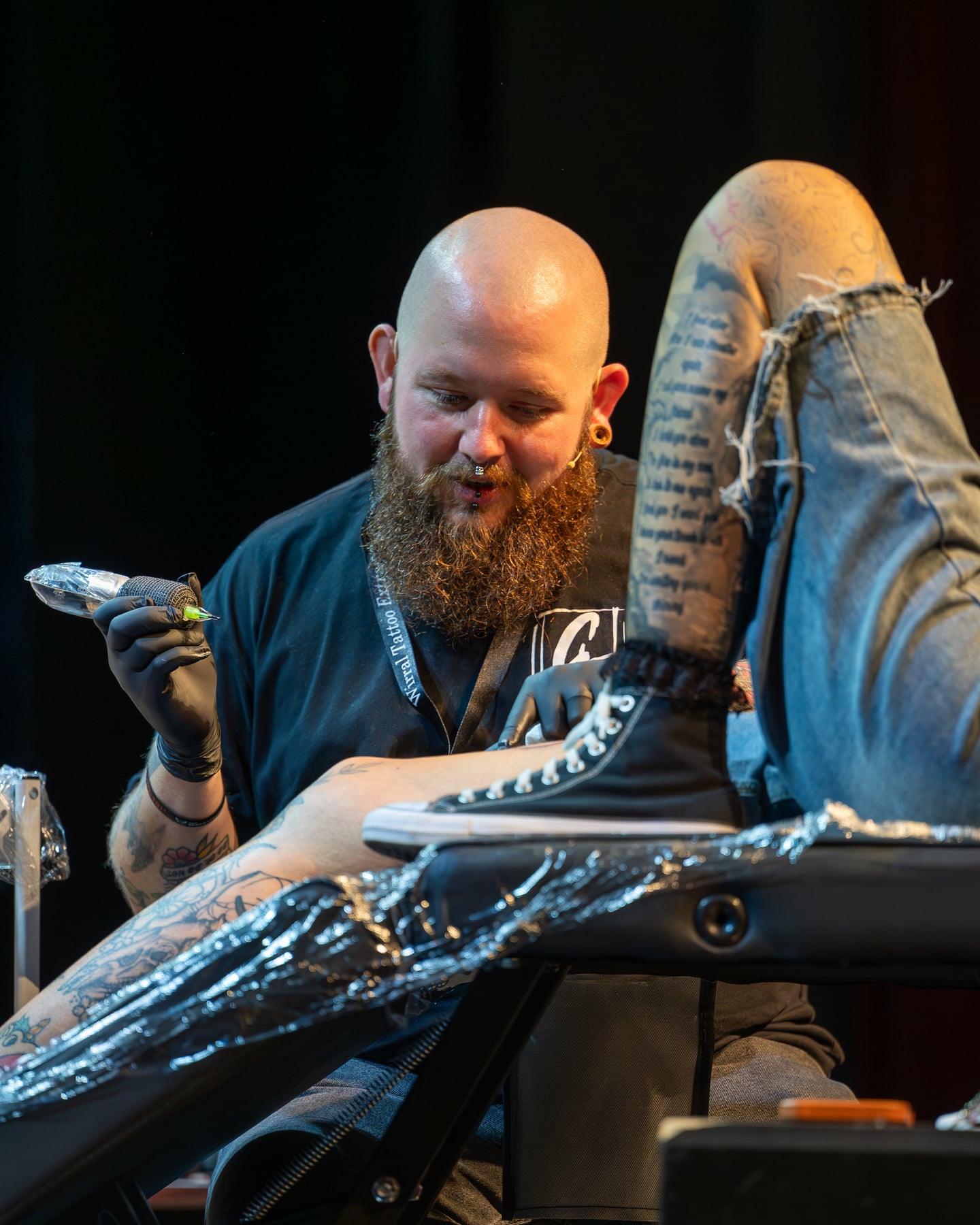 A bearded tattoo artist working beside a client's raised legs.