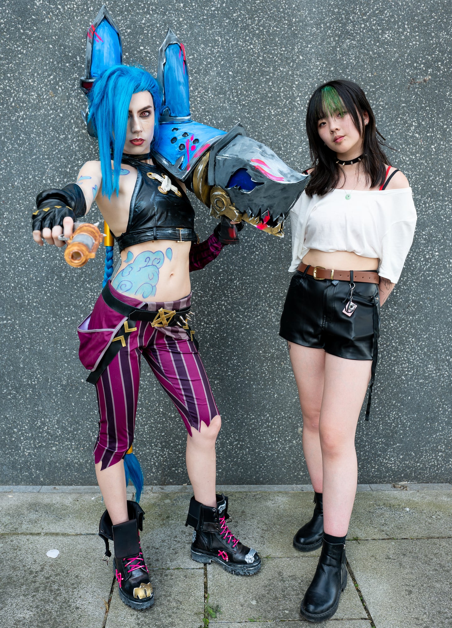 Two cosplayers, one with long blue hair, posing against a stone wall.