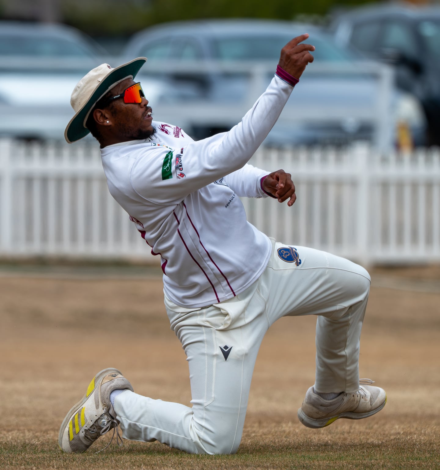 A cricket fielder in a sun hat reaching upwards from one knee.