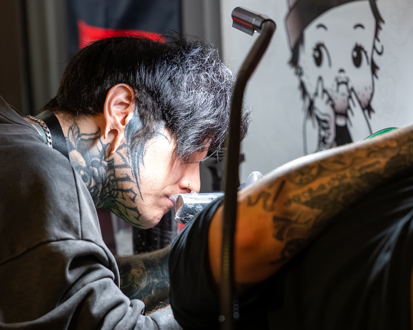 A dark-haired tattoo artist leaning towards a client's arm.
