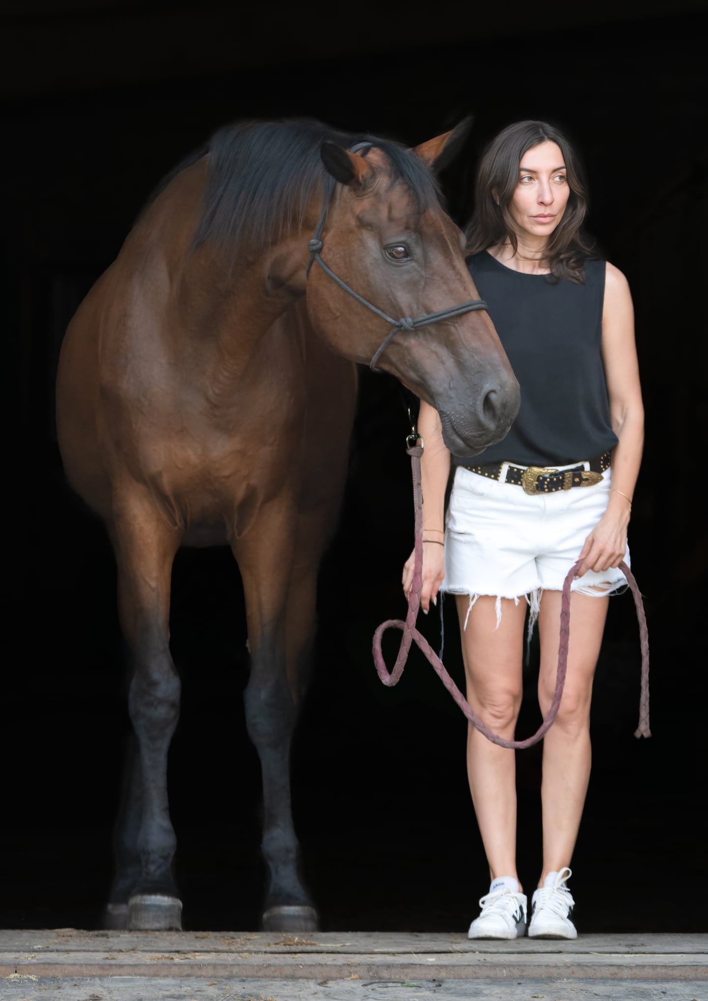 A woman holding a lead rope beside a bay horse against a dark background.