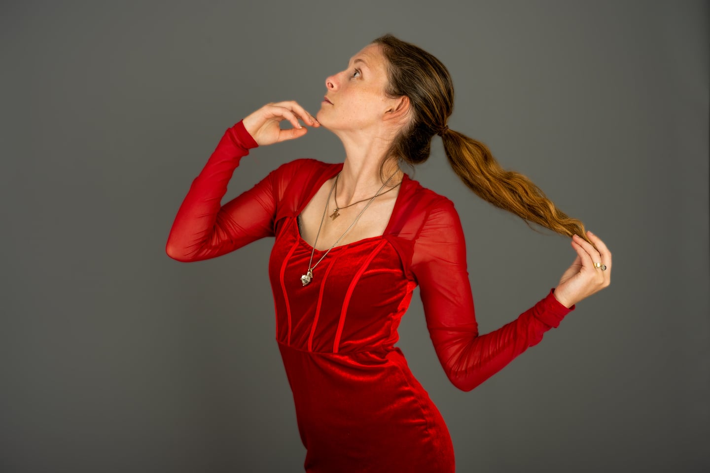 A model in a red dress holding her hair against a grey studio backdrop.