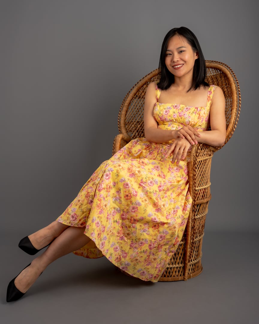 A model in a yellow floral dress seated in a wicker chair in the studio.