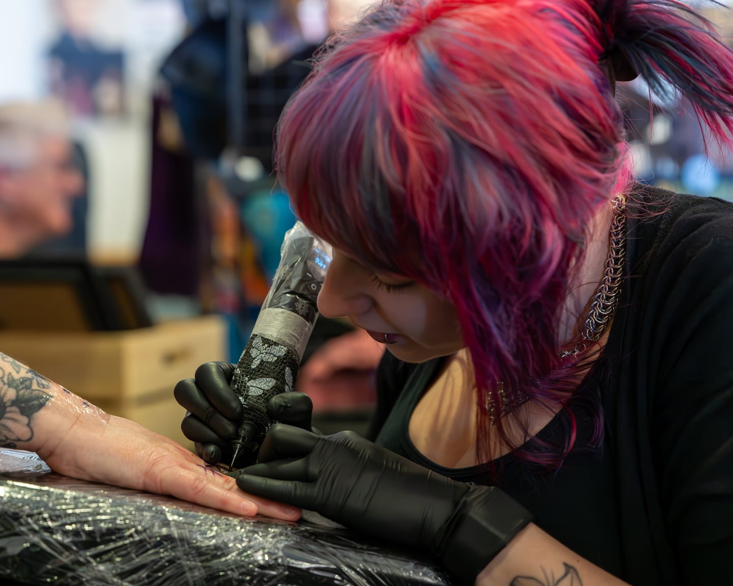 A pink-haired tattoo artist leaning over a client's arm.