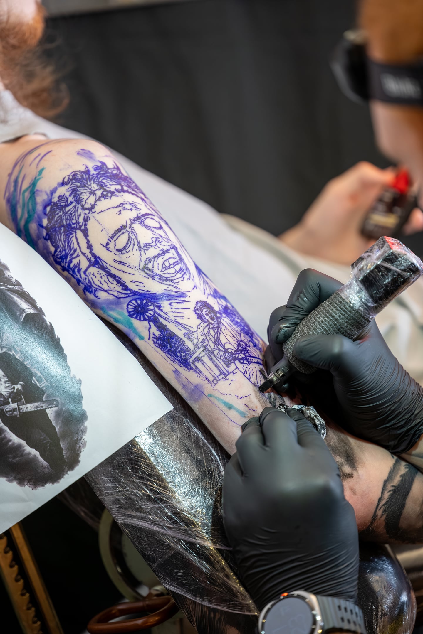 A gloved tattoo artist working over a purple stencil on a client's leg.