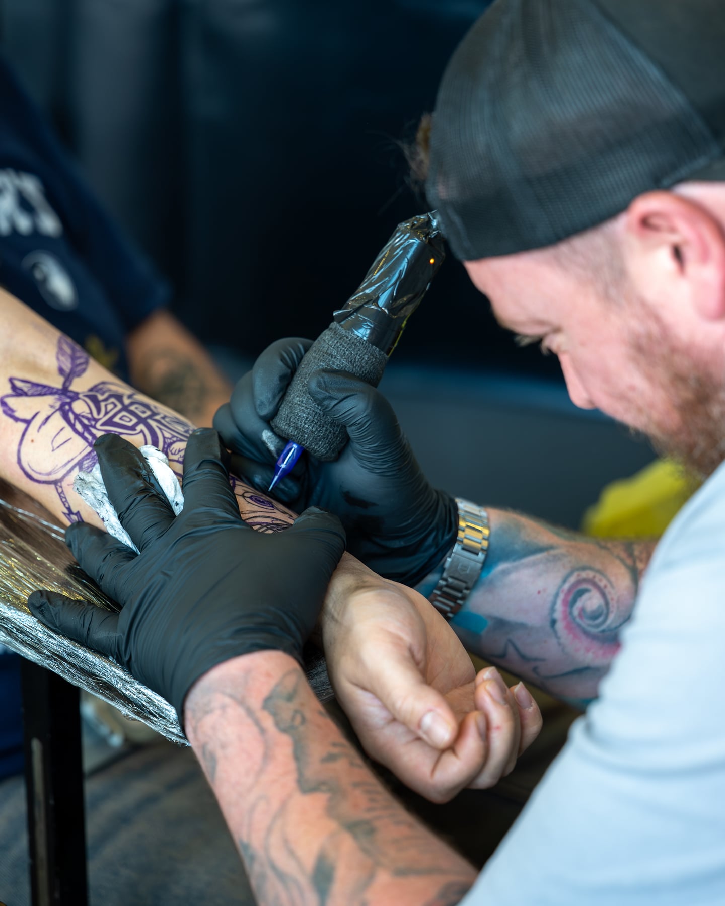 A tattoo artist in a backwards cap concentrating on a client's tattoo.