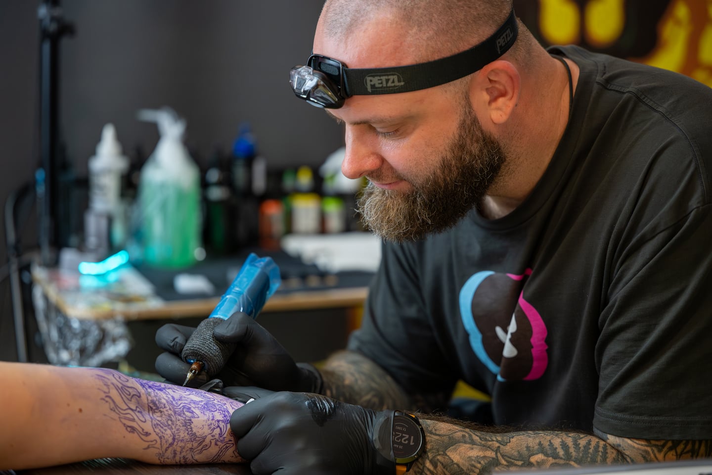 A tattoo artist wearing a headlamp concentrating on a client's arm.