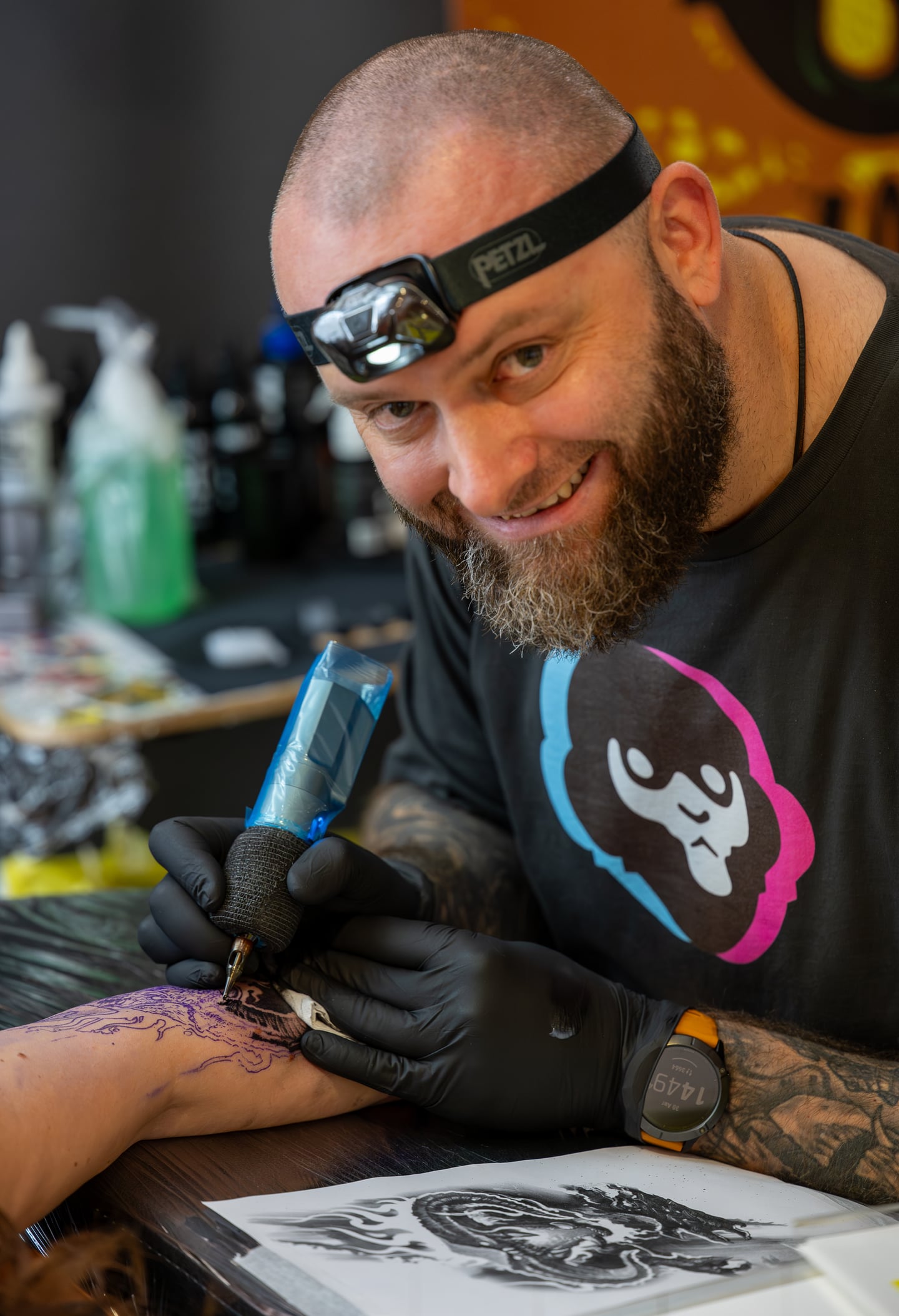 A smiling tattoo artist wearing a headlamp and black gloves.