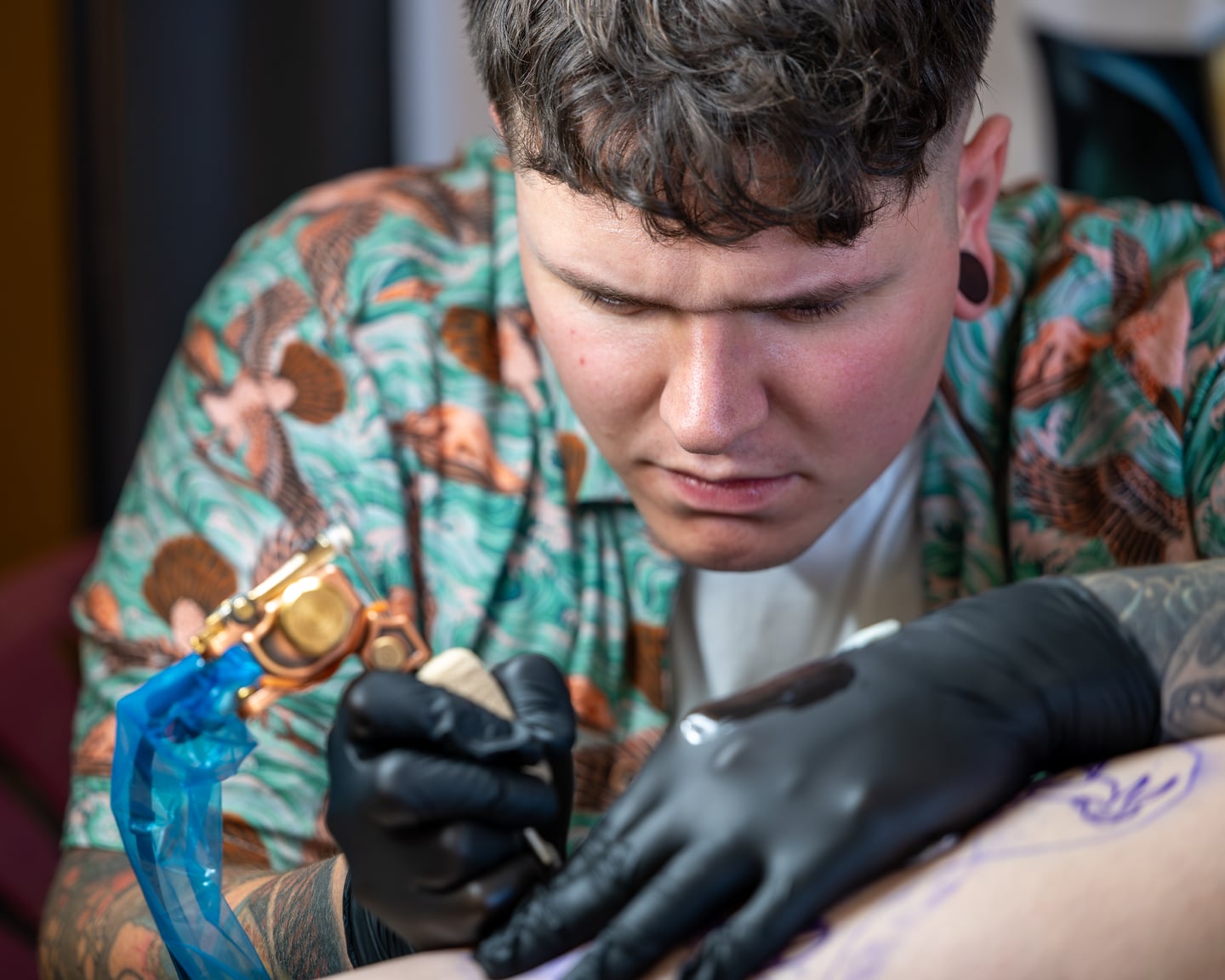 A tattoo artist in a patterned shirt working with black gloves.