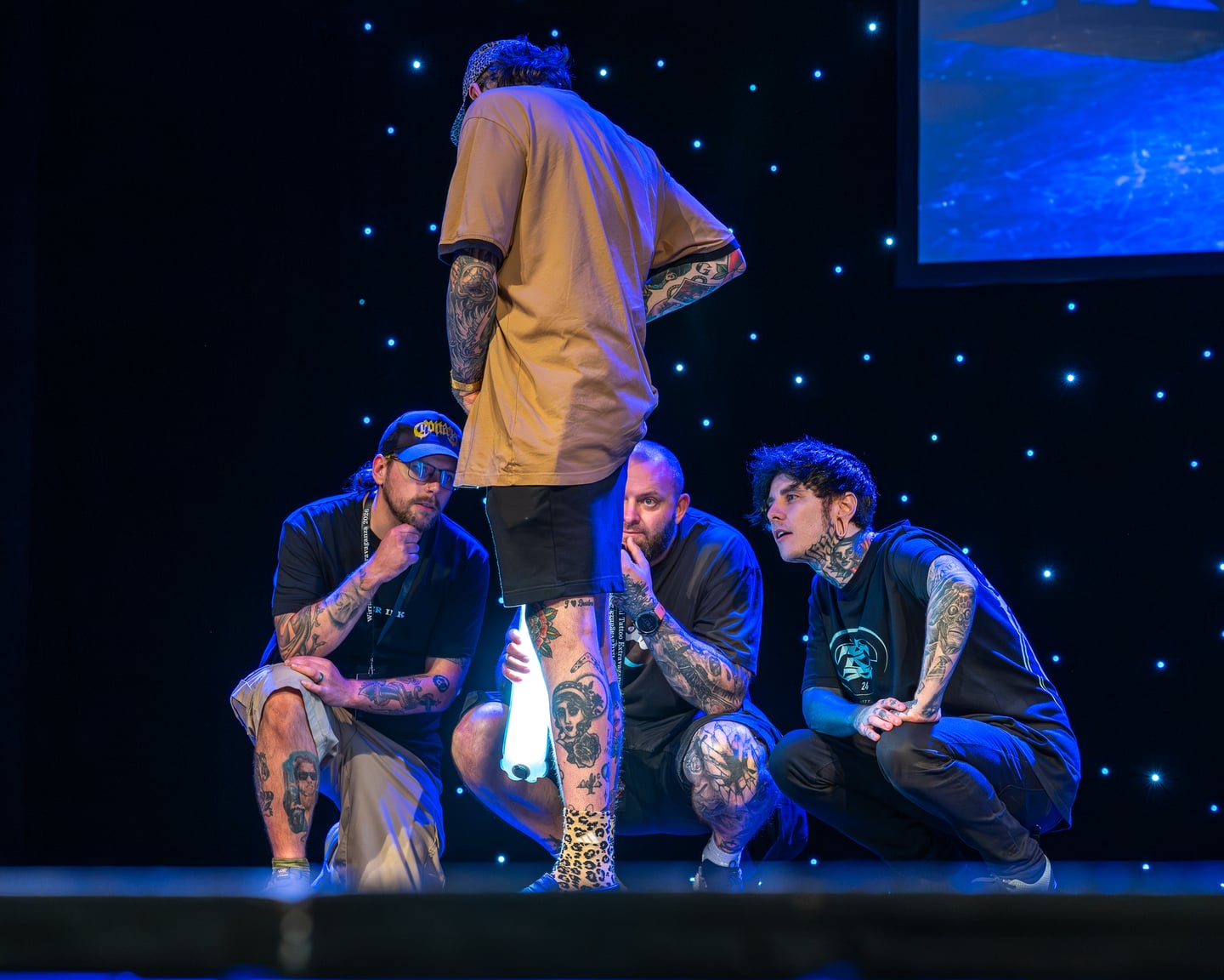 Tattoo event judges examining a participant's leg tattoos on stage.