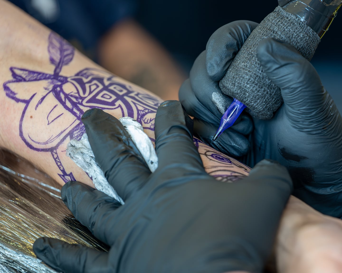 Close-up of gloved hands and a tattoo machine following a purple stencil.